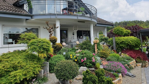 Une maison à deux étages dotée d’un balcon, entourée d’un jardin bien entretenu où poussent diverses plantes et fleurs.