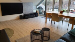Un salon moderne doté d’un téléviseur à écran plat, d’un coin repas avec une table et des chaises, et d’une grande fenêtre offrant une vue sur l’extérieur.