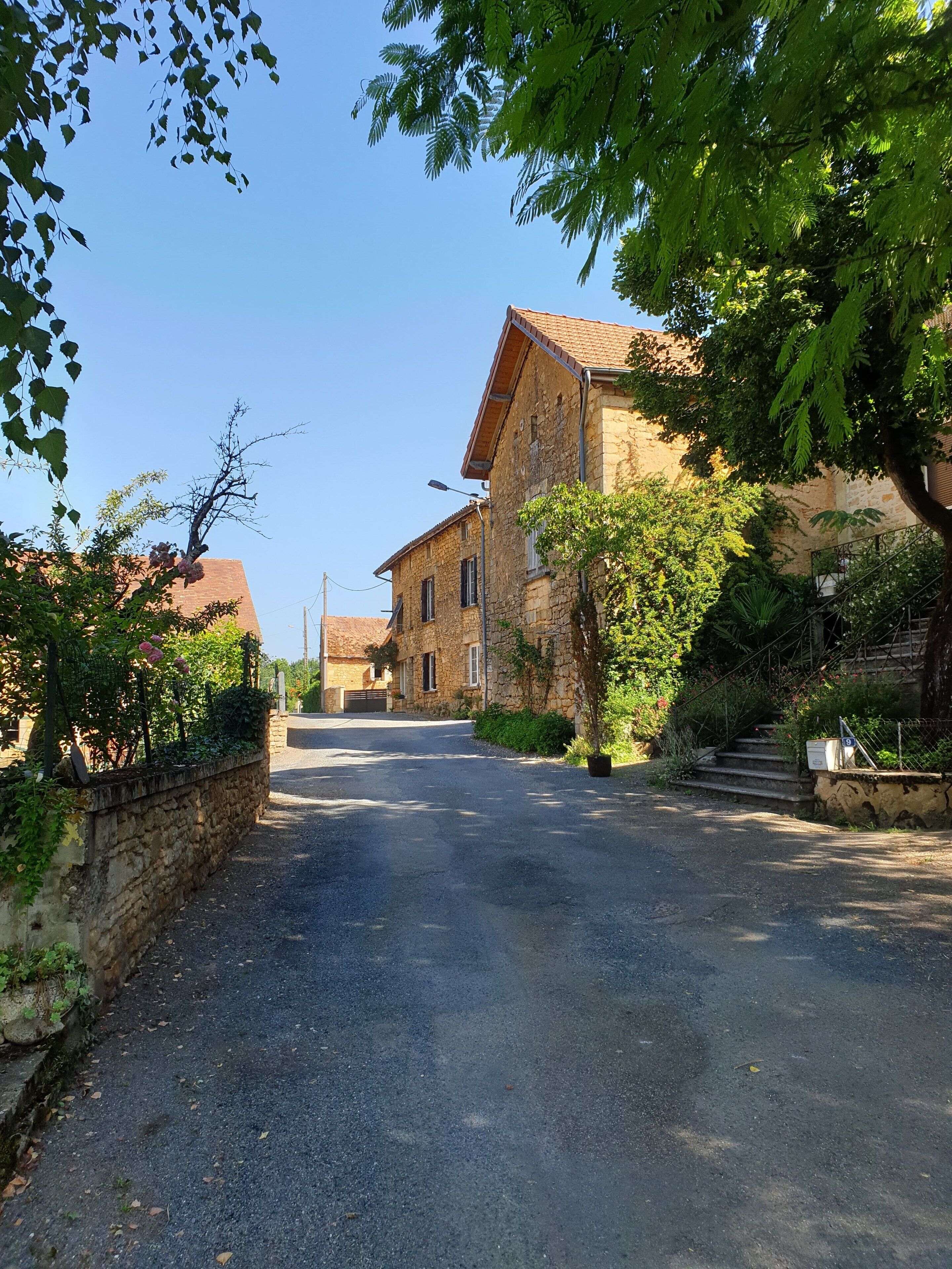 A quiet street with stone buildings, a paved road, and lush greenery.