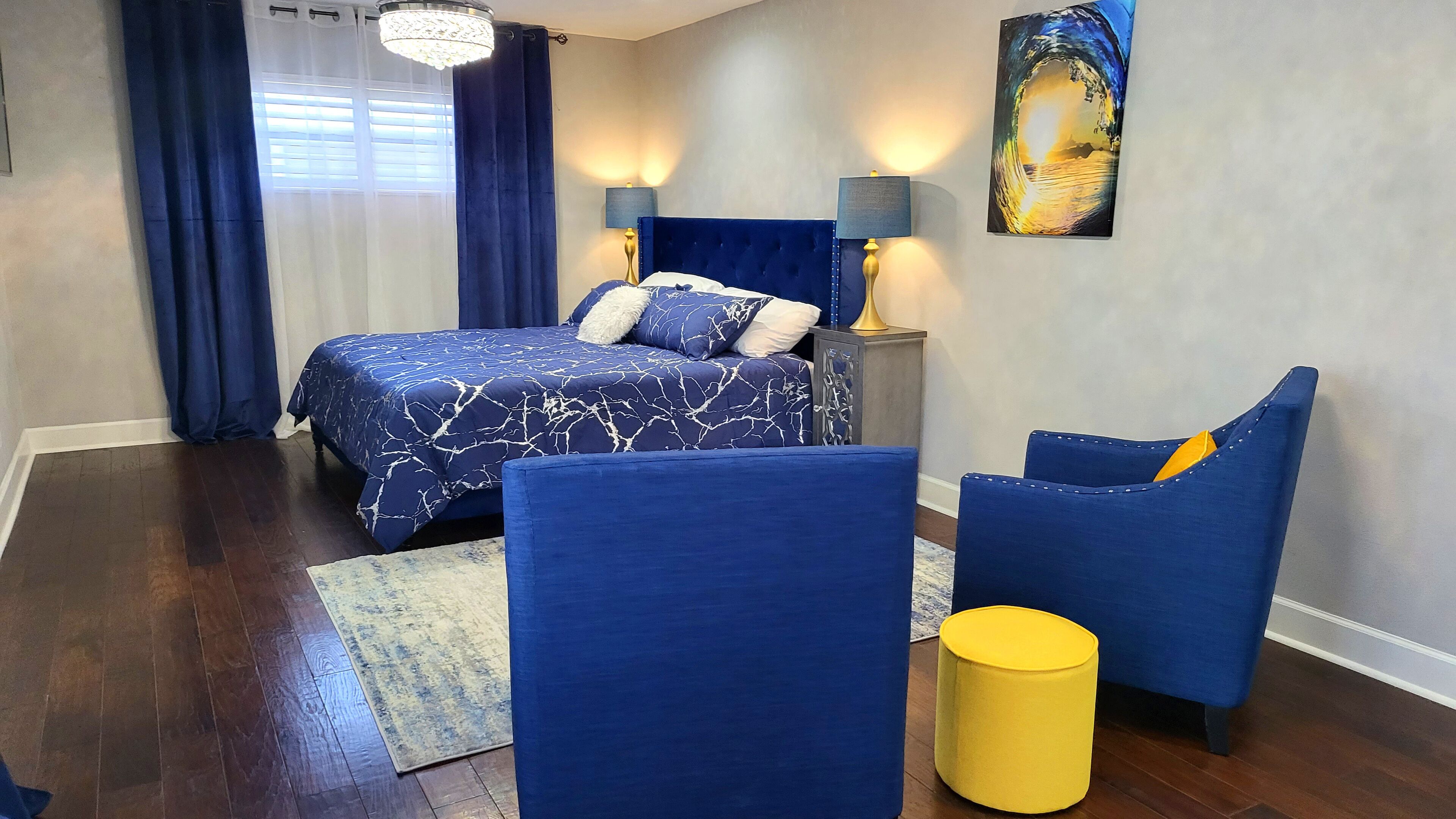 A bedroom with a bed, two chairs, a nightstand, a lamp, and a painting on the wall.