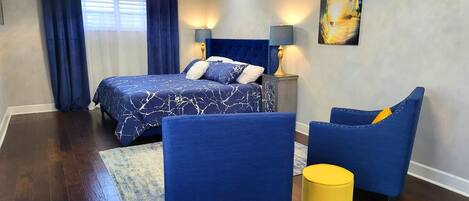 A bedroom with a bed, two chairs, a nightstand, a lamp, and a painting on the wall.