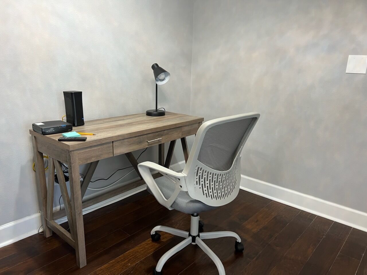 Un escritorio de madera con una computadora, una lámpara y una silla en una habitación con pared gris y suelo de madera.