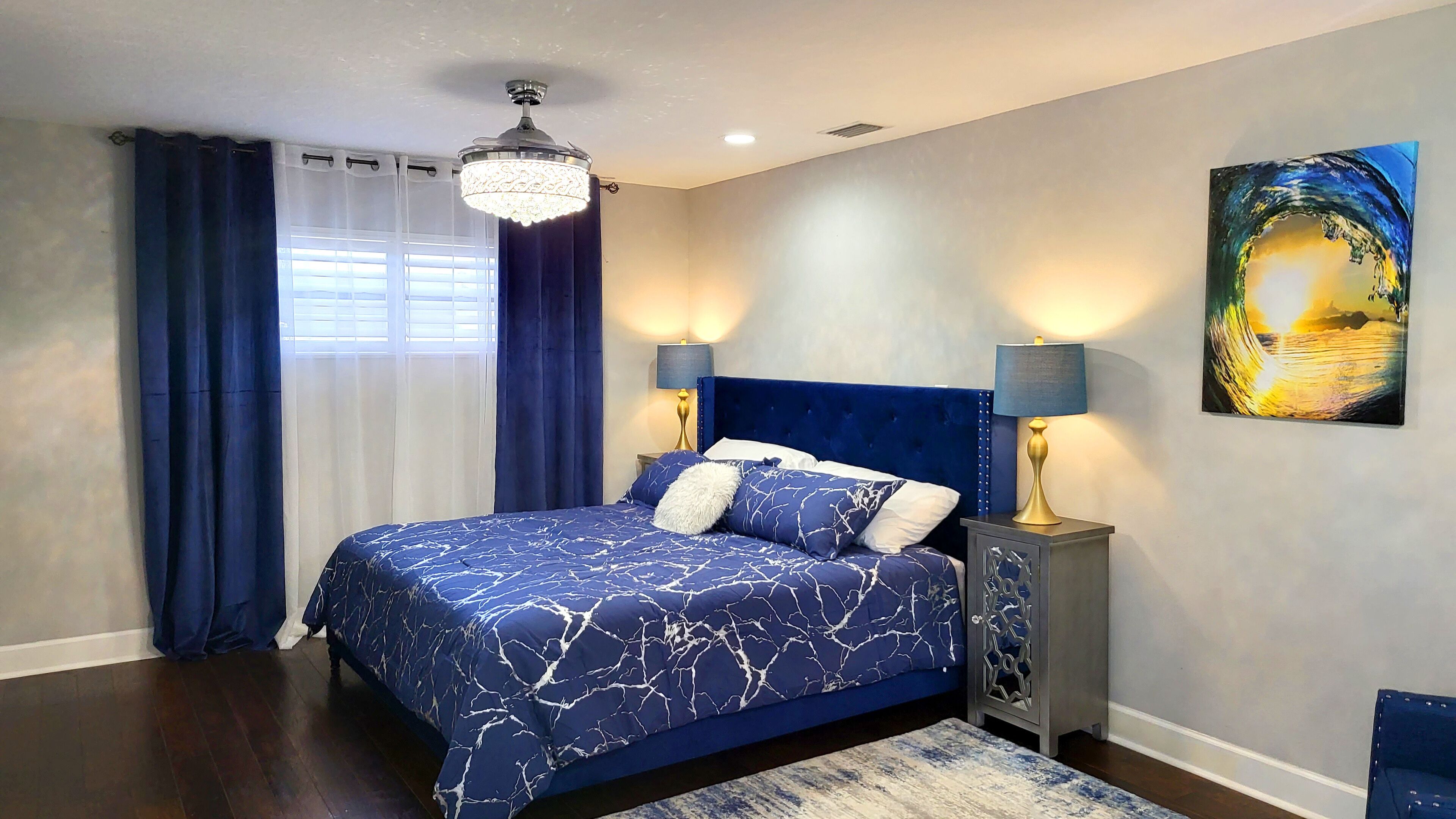 A bedroom with a bed, curtains, a chandelier, a nightstand, and a wall painting.