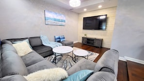 Un soggiorno moderno con un divano modulare grigio, due tavolini da caffè in marmo bianco, una TV a schermo piatto montata su una parete in legno e un quadro raffigurante una spiaggia.