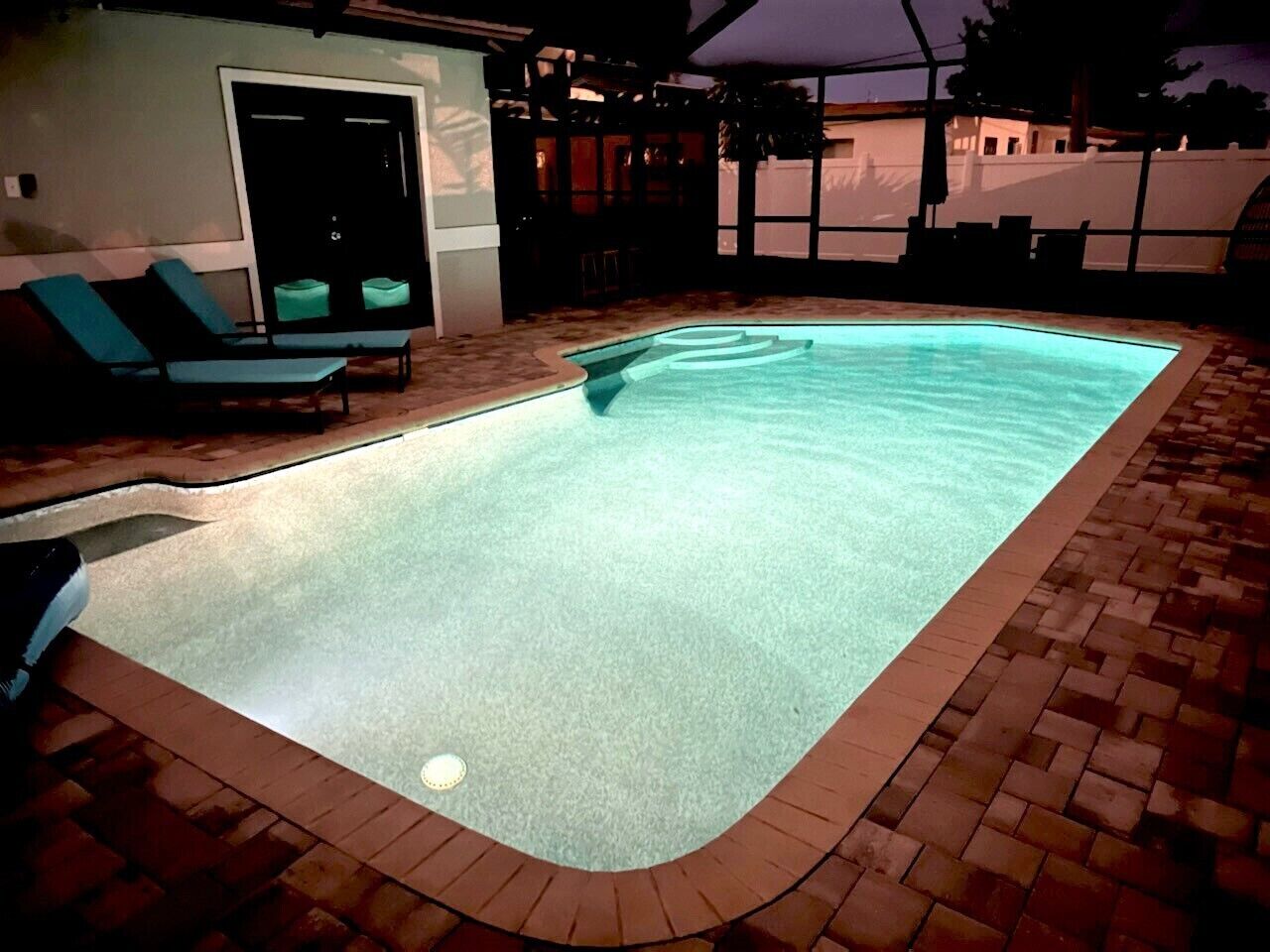 Una piscina con borde de azulejos y un pequeño jacuzzi circular. Se ven sillas de descanso y una zona cubierta al fondo.