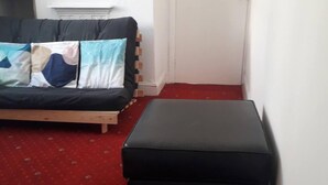 Een kleine kamer met een bed, een bankje en een rood tapijt.