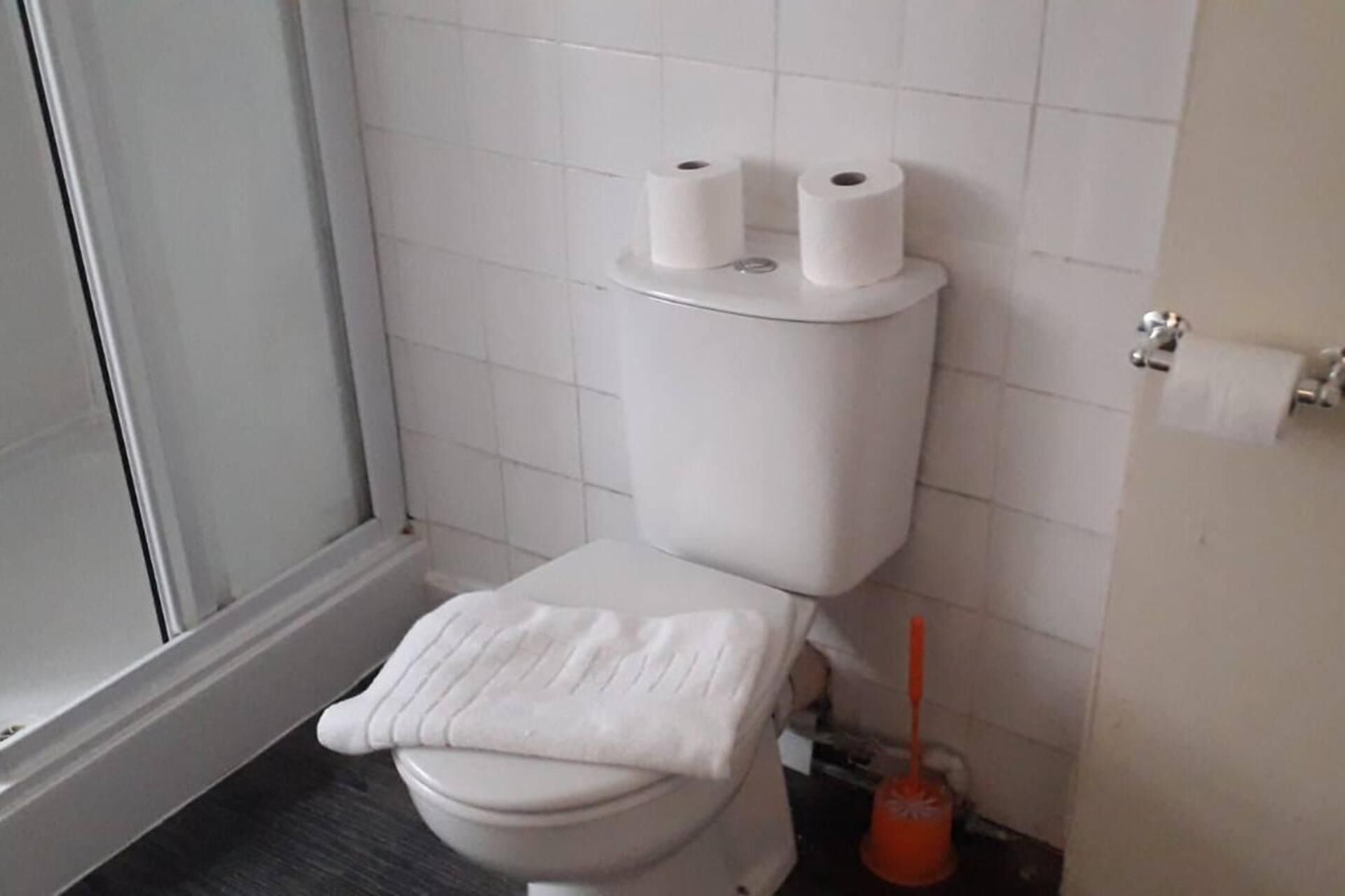 Un baño con un inodoro, dos rollos de papel higiénico en el tanque, una toalla blanca sobre el asiento y un desatascador en el suelo.