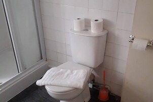 Un baño con un inodoro, dos rollos de papel higiénico en el tanque, una toalla blanca sobre el asiento y un desatascador en el suelo.