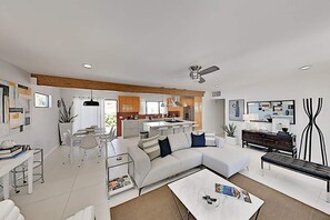 Un salon moderne avec un canapé blanc, une table basse et un coin repas comprenant une table et des chaises blanches. On y trouve également une cuisine avec un grand îlot central et un ventilateur de plafond.