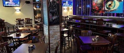 Un interior de restaurante con mesas y sillas de madera, un menú en una pizarra y una pantalla de televisión grande mostrando un noticiero.