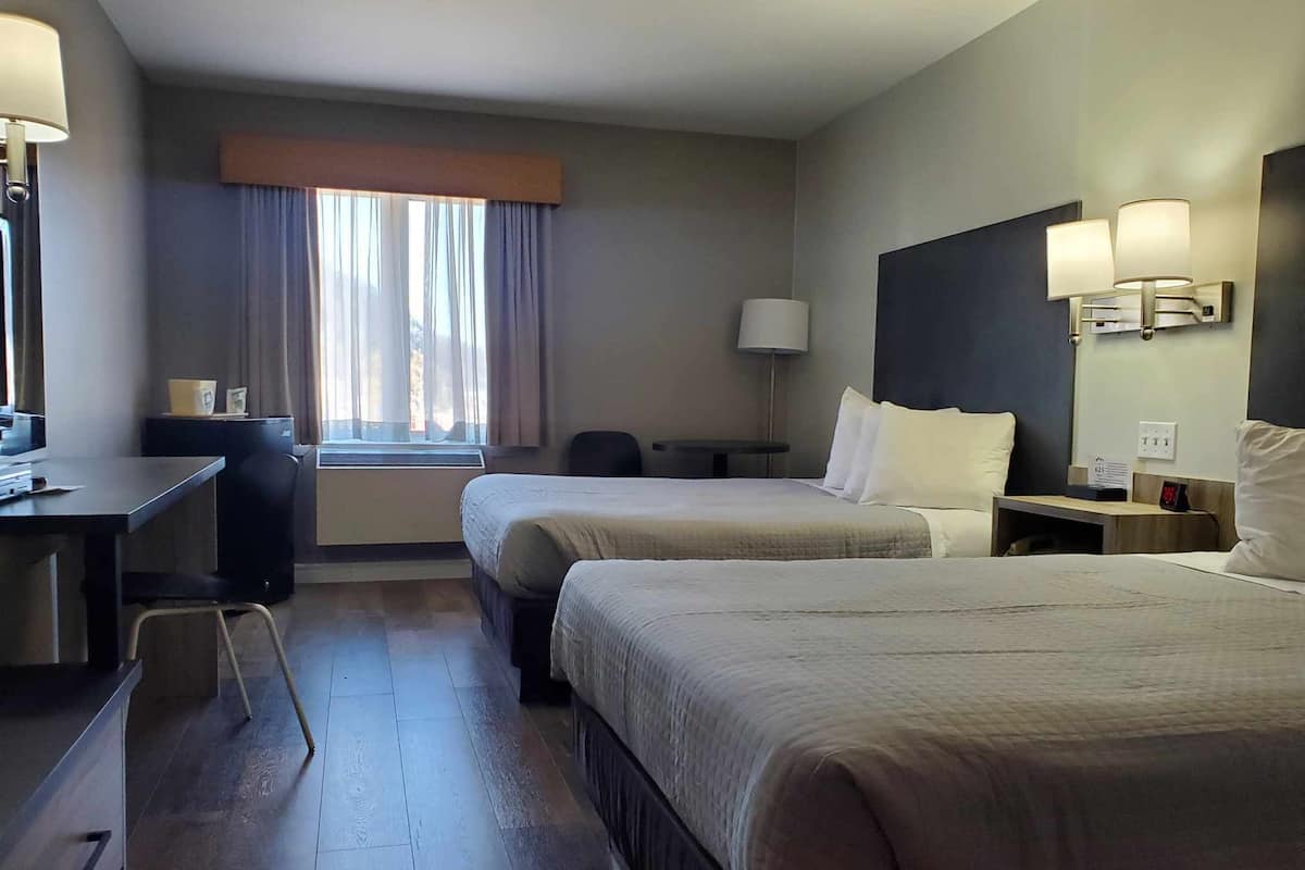 Habitación de hotel con dos camas, un escritorio, una silla, ventana con cortinas y una lámpara fijada a la pared.