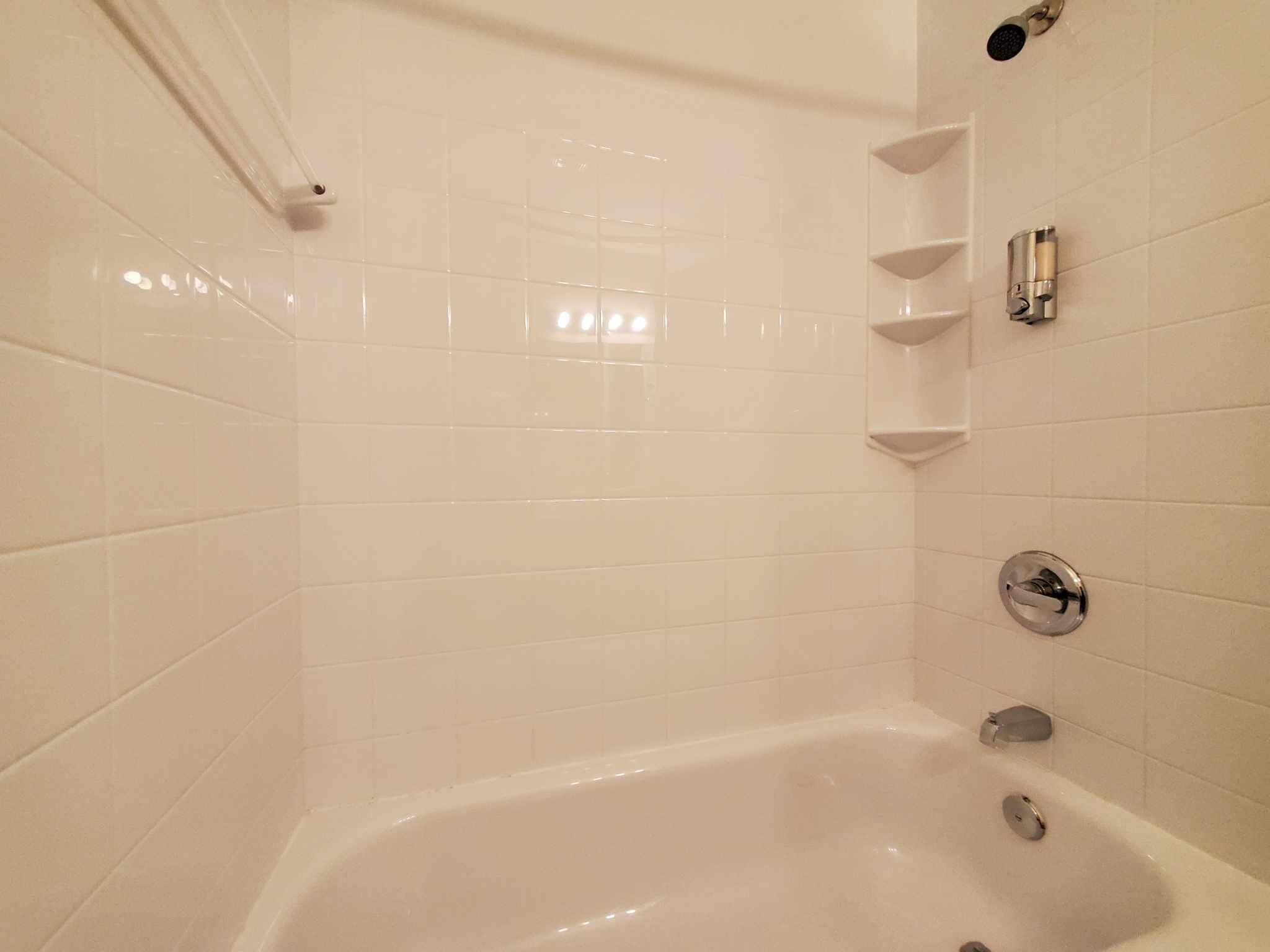 Une salle de bain avec des murs recouverts de carreaux blancs, une baignoire et une pomme de douche.