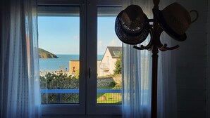 A view through a window with a curtain, overlooking a coastal area with a house and a hill.