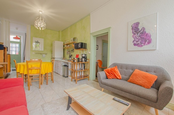 Una sala de estar con un sofá gris, un sofá rojo y una mesa de centro de madera. Se observa una zona de cocina con un refrigerador, microondas y diversos utensilios. También se ve una mesa de comedor con manteles amarillos y sillas.