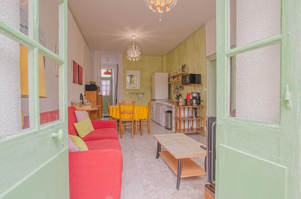 Un salón acogedor con un sofá rojo, una mesa de comedor de madera y una zona de cocina con una cafetera y estanterías.