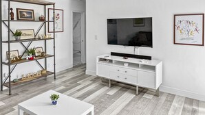 Una sala de estar moderna con un mueble blanco para el televisor, una mesita blanca y una estantería de madera con varios objetos decorativos.