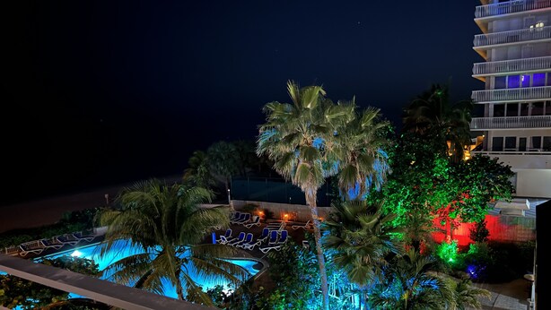 A nighttime view of a resort with a pool area, palm trees, and a building with balconies. - Beautiful Oceanview Modern Hotel Room W/ Balcony (Fort Lauderdale)