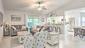 A spacious living area with a sofa, coffee table, and patterned armchair. A ceiling fan and a dining area with chairs and a table are also visible.