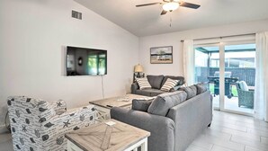 A living room with a ceiling fan, a flat-screen TV, a grey sofa, patterned armchairs, and a dining table.