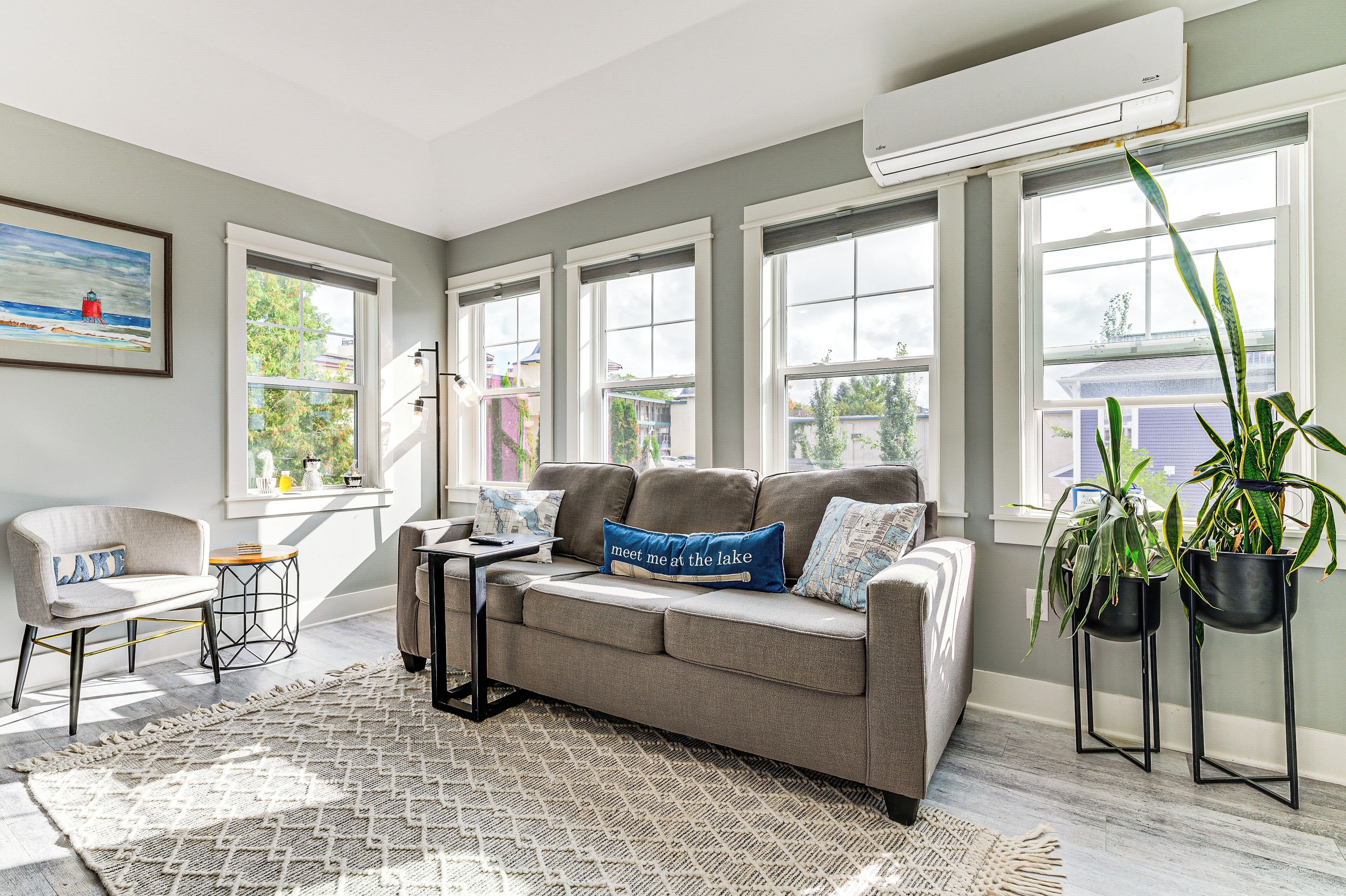 A living room with a grey sofa, a small table, a chair, a patterned rug, and large windows with a view of the outdoors.