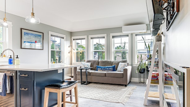 Eine moderne Küche mit einer zentralen Kochinsel, ein Essbereich mit grauem Sofa und Blick ins Freie durch große Fenster.