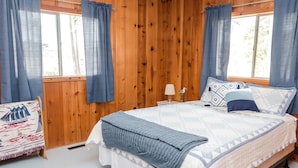 Quarto com painéis de madeira, uma cama, cortinas, uma luminária e uma cadeira.