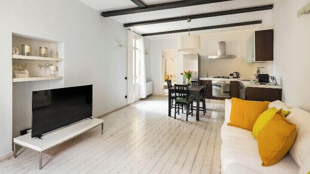 A modern living room with a flat-screen TV, a dining area with a table and chairs, a sofa with yellow cushions, and a kitchen in the background.