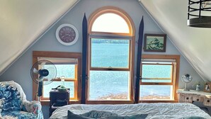 Een slaapkamer met een groot bed, twee ramen met uitzicht op zee, een klok aan de muur en een ventilator op een nachtkastje.