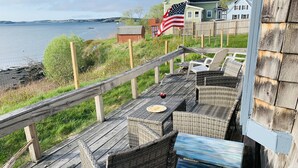 Een houten terras met rieten meubilair, met uitzicht op een waterpartij, een huis met vlag en een houten omheining.
