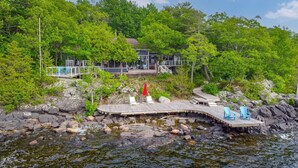 A wooden dock extending into a rocky shoreline with a small bridge connecting to a house surrounded by lush greenery. - Beautiful Luxury Lake Front Cottage Georgian Bay (Georgian Bay)