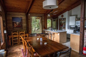 Un coin repas rustique en bois, avec une grande table et des chaises, une cuisine dotée de meubles blancs et une vue sur l’extérieur par une fenêtre.