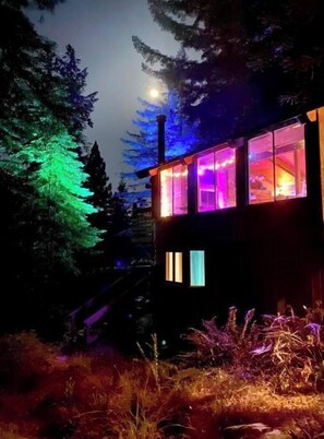 Un chalet doté de grandes fenêtres illuminées par des lumières colorées, entouré d’arbres et avec une montagne en arrière-plan.