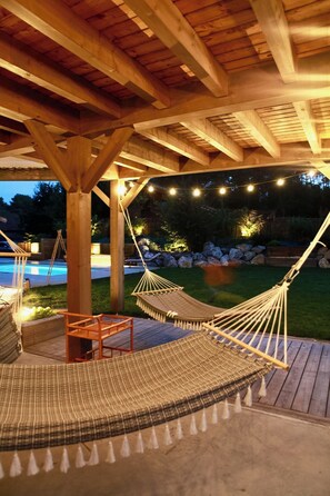 Une terrasse en bois avec un hamac et une table, entourée d’une piscine et d’un jardin.