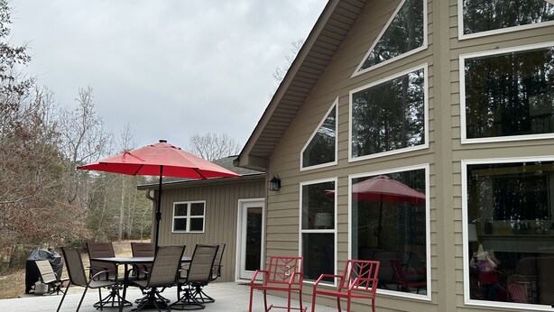 An outdoor deck with a dining table and chairs, a red umbrella, and a large window overlooking a wooded area. - Lake Hartwell on McKinney Point, near Clemson! (Westminster)