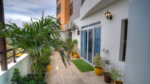 Un balcon étroit agrémenté de plantes en pot, un petit espace recouvert de gazon artificiel et un immeuble à la façade blanche et aux grandes fenêtres.