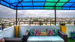 Un balcon donnant sur un paysage urbain, un canapé agrémenté de coussins colorés et un auvent bleu et vert.