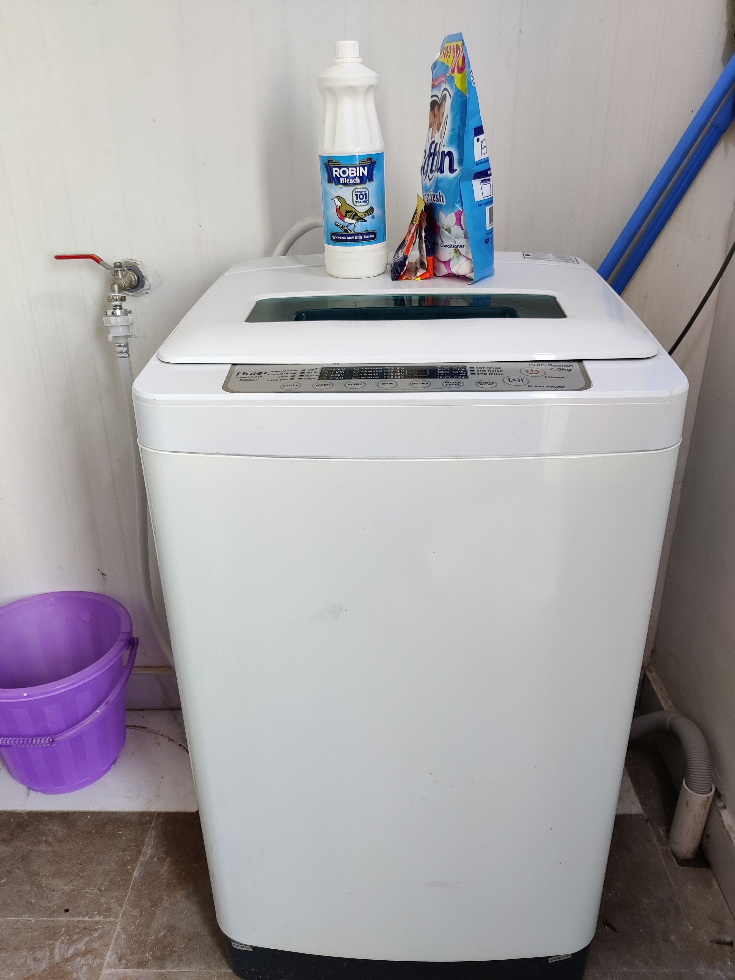 Une machine à laver blanche, dotée d’un panneau de commande, un seau violet et des produits de nettoyage sont posés sur le dessus.
