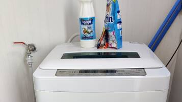 Une machine à laver blanche, dotée d’un panneau de commande, un seau violet et des produits de nettoyage sont posés sur le dessus.