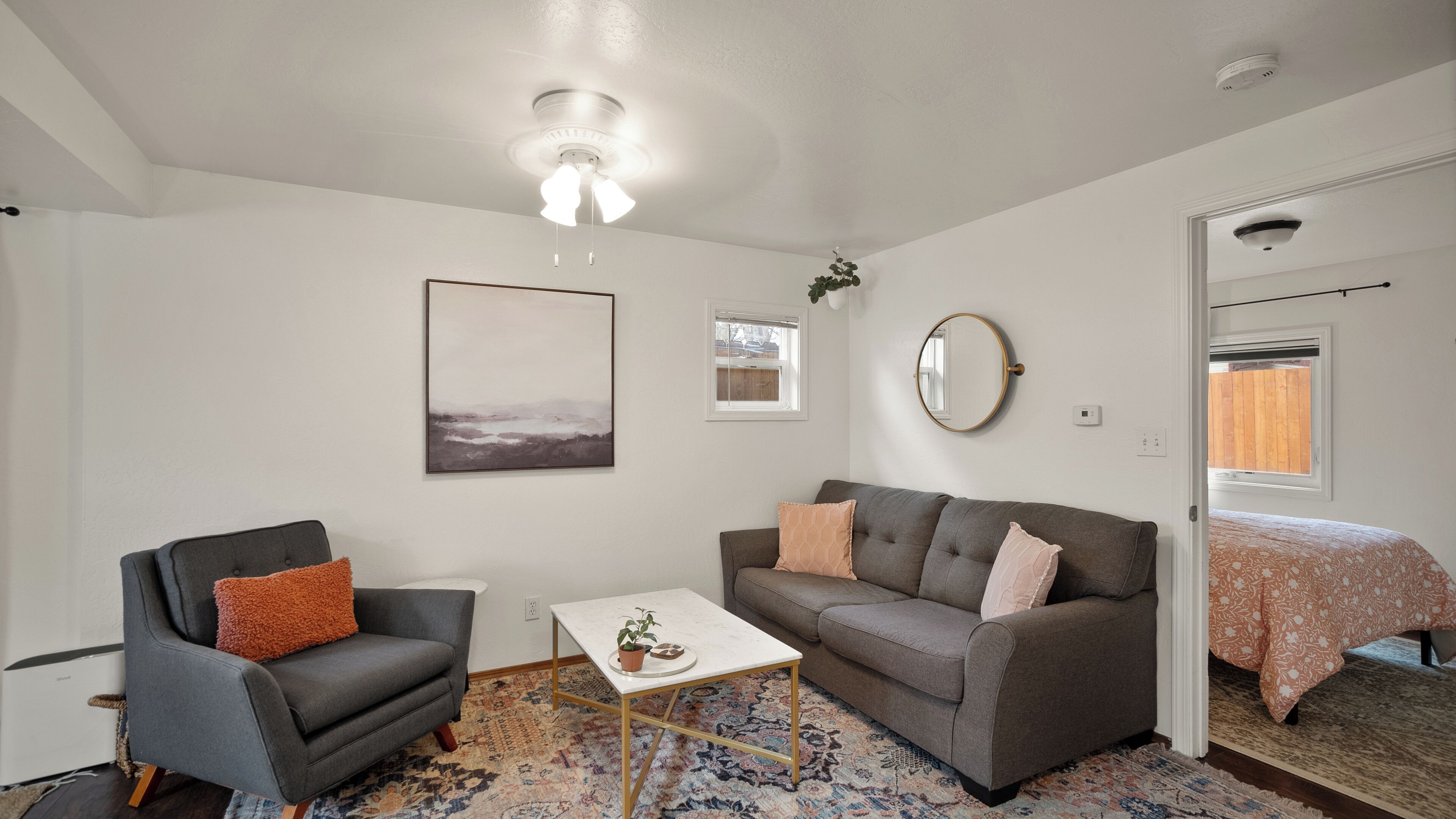 Una sala de estar moderna con un sofá gris, un sillón más pequeño, una mesa de centro y una alfombra estampada. También se observa un ventilador de techo y un espejo adosado a la pared.