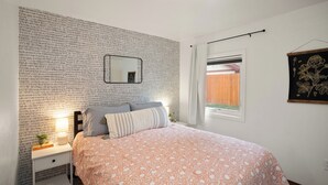 A bedroom with a patterned wall, a bed with a floral bedspread, a bedside table with a lamp, and a window with a view of a wooden fence. - Downtown CDA Tiny House-Walk to the Lake (Coeur d'Alene)