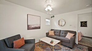 A living room with a grey sofa, a smaller grey armchair, a coffee table, a floor lamp, a ceiling fan, a framed picture, a mirror, and a window. - Downtown CDA Tiny House-Walk to the Lake (Coeur d'Alene)