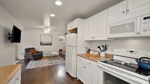 A modern kitchen with white cabinets, a microwave, a stove, and a refrigerator. There is a dining area with a table and chairs, and a living room with a sofa and a TV. - Downtown CDA Tiny House-Walk to the Lake (Coeur d'Alene)