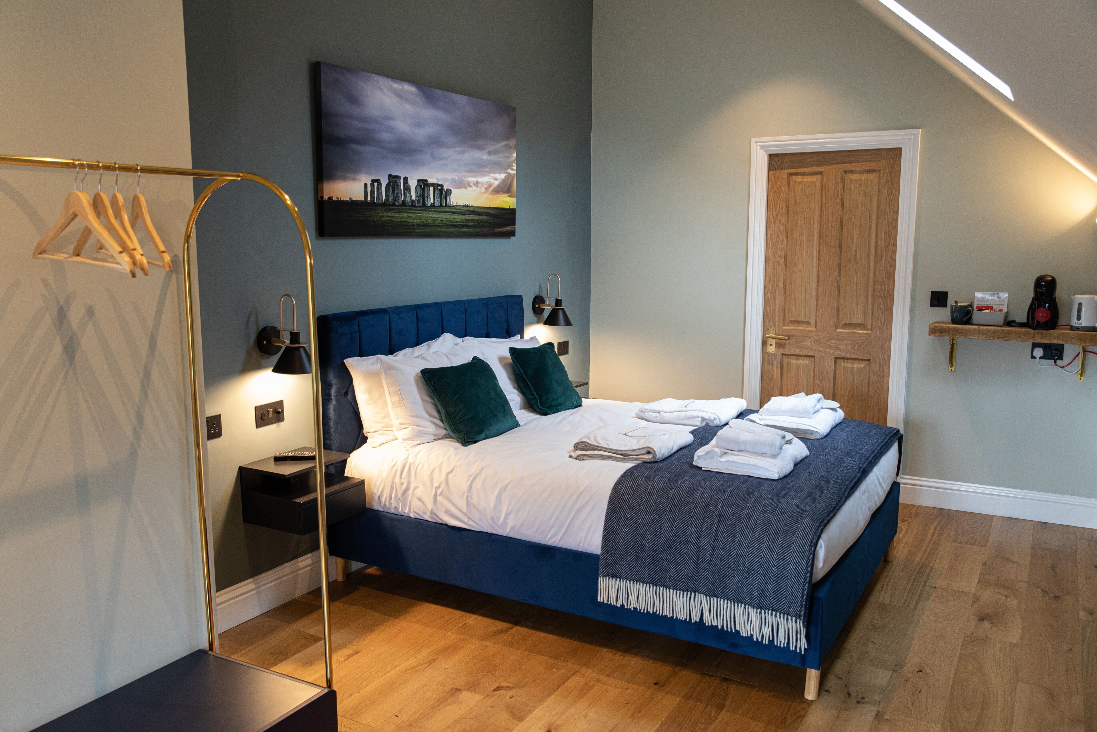 A neatly made bed with a blue headboard, a bedside table with a lamp, a wooden door, and a wall-mounted shelf with items.