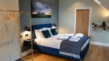 Una cama bien hecha con cabecera azul, una mesita de noche con lámpara, una puerta de madera y un estante con objetos.