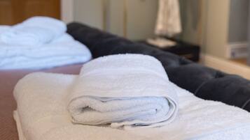 A neatly made bed with folded white towels, a wooden door, and a robe hanging on a rack in a room with light-colored walls.