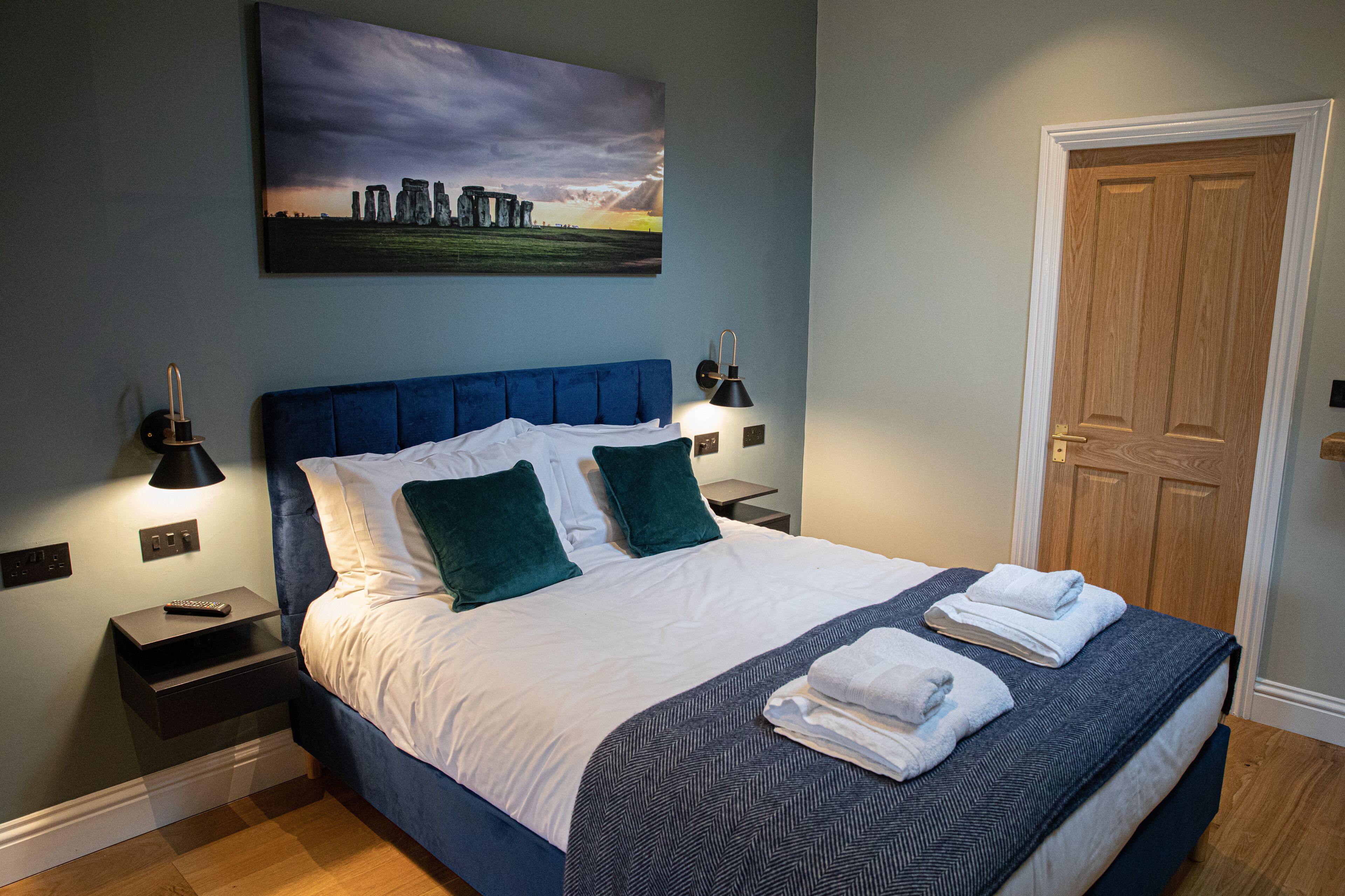 A neatly made bed with a blue headboard, two bedside lamps, and a painting of Stonehenge on the wall.