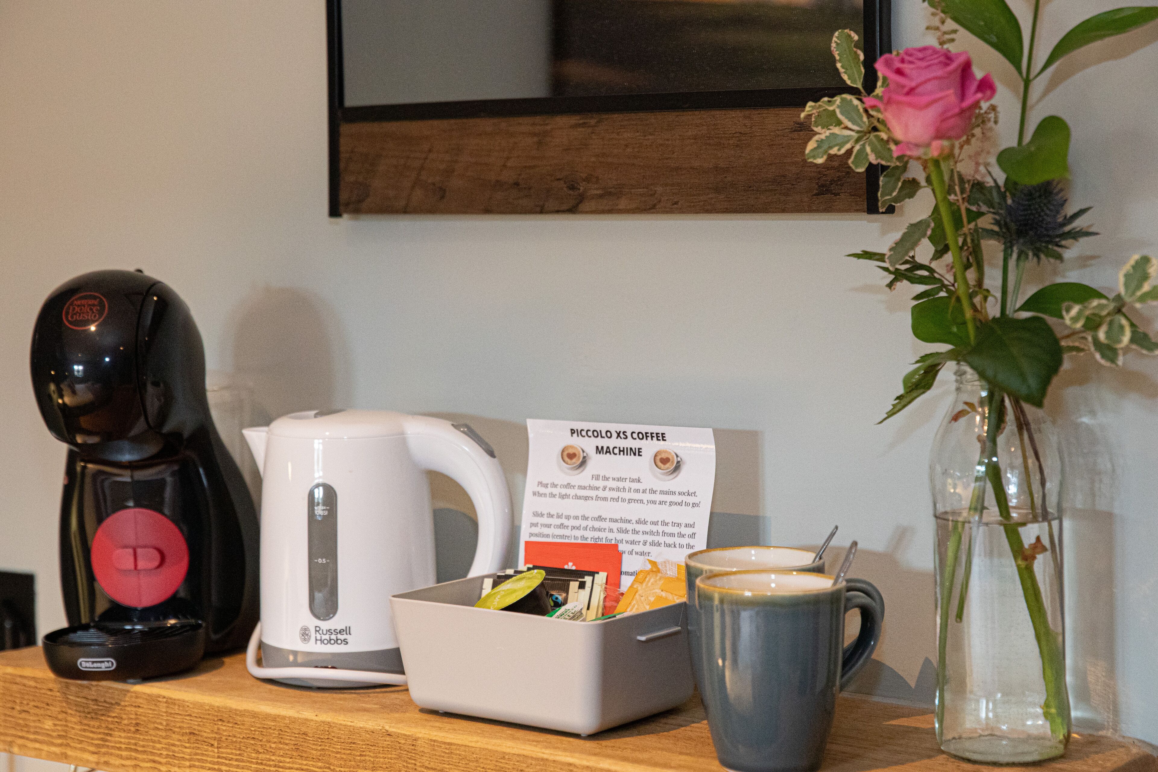 Uma máquina de café, uma chaleira, uma caneca, um vaso de flores e uma placa com instruções.