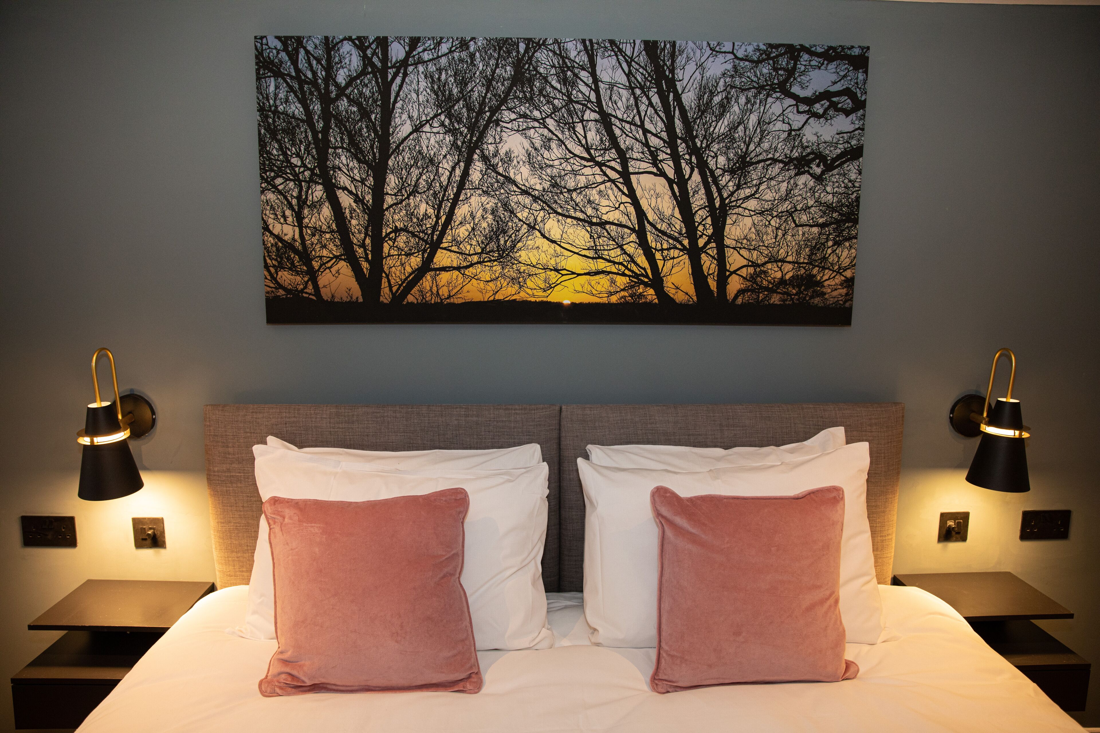 A hotel room with a large bed, two nightstands, and a wall-mounted painting of trees at sunset.