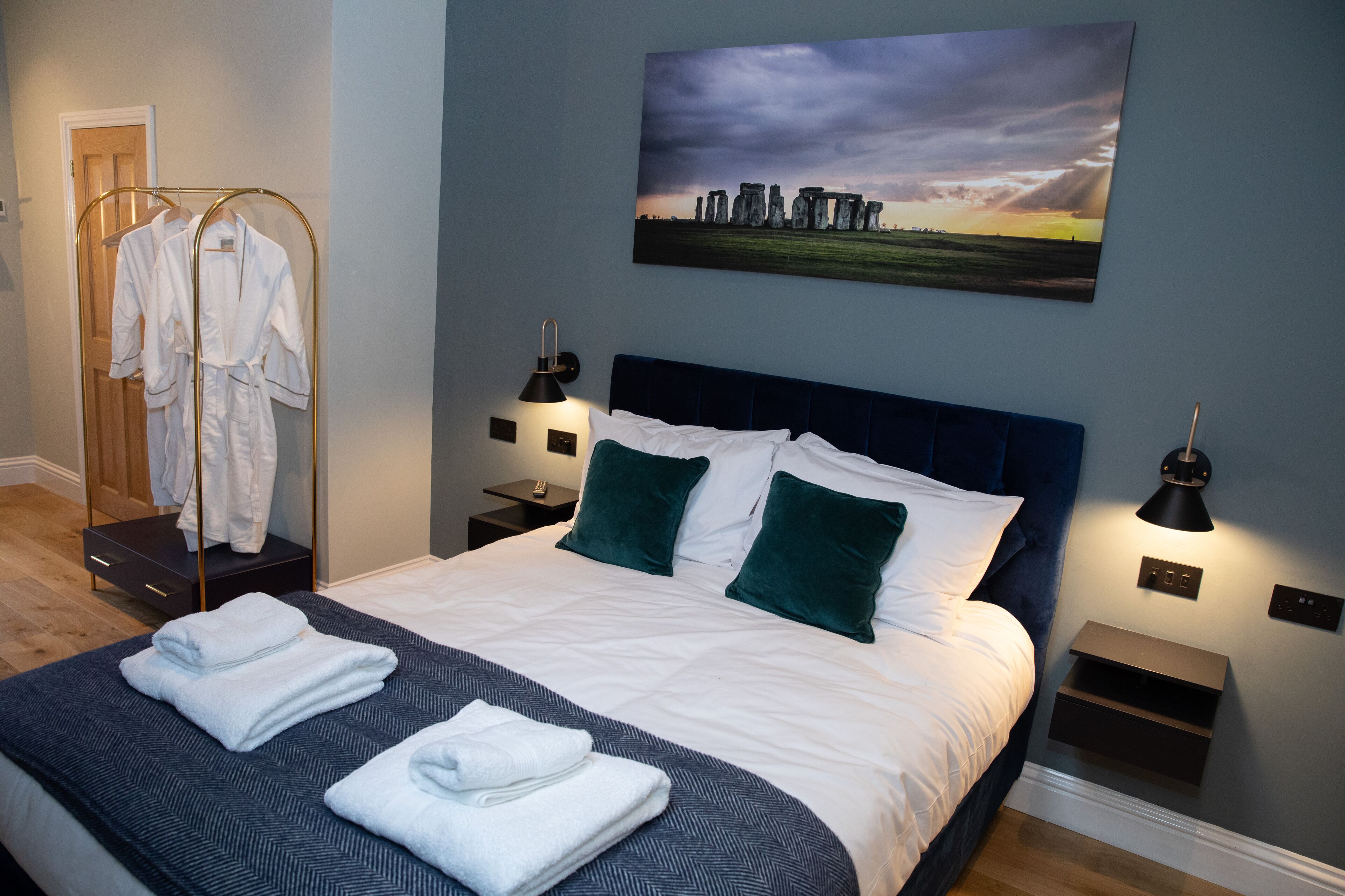 A neatly made bed with white linens and green pillows, a bedside table with a lamp, and a wall-mounted painting of Stonehenge.