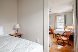 Una habitación de hotel con una cama, una mesita de noche de madera, un sillón mecedora y una ventana con cortinas.